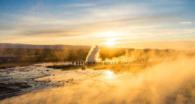 Menigten kijken naar een hoge geiser die uitbarst te midden van gouden stoom bij zonsondergang over een reflecterende natte vlakte.