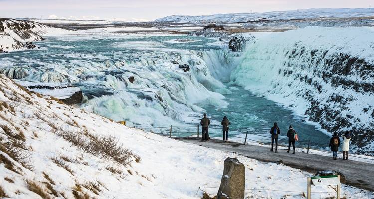Besneeuwde kliffen omlijsten een gedeeltelijk bevroren Gullfoss waterval terwijl een handvol bezoekers bij de reling staat.