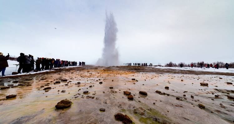 Winterse scène van een geiser die hoog opspuit in een bleke lucht terwijl twee rijen ingepakte toeristen toekijken vanaf de zijkanten van met mineralen bevlekte grond.