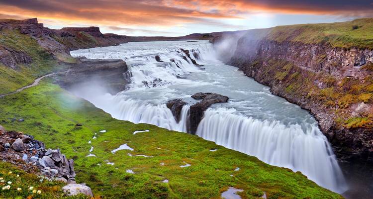 Levendige zonsondergangkleuren verlichten de mistige Gullfoss-waterval die naast heldere groene bemoste oevers naar beneden stroomt.