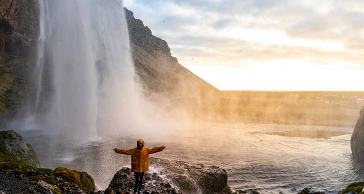 Blije reiziger in een gele jas spreidt de armen onder de nevel van de Seljalandsfoss waterval bij zonsopgang.