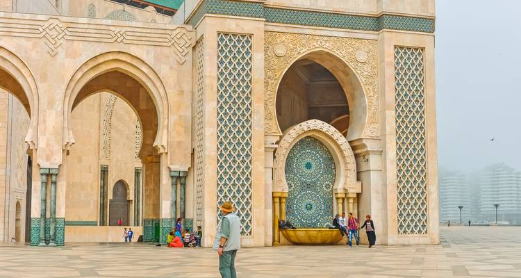 Besucher, die über den gefliesten Innenhof der Hassan-II-Moschee mit kunstvollen Bögen an einem nebligen Morgen gehen.