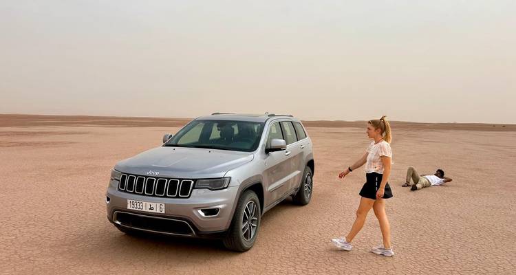 SUV geparkt auf weiter trockener Wüstenpfanne, während eine Frau läuft und ein Mann am Boden liegt und posiert.