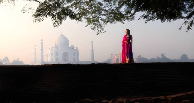 Woman in vibrant traditional dress standing on a wall with the Taj Mahal glowing in morning mist behind her.