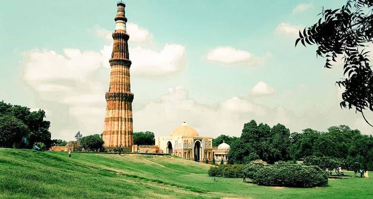 Tall brick minaret of Qutub Minar rising above green lawns and scattered trees beneath a partly cloudy sky.