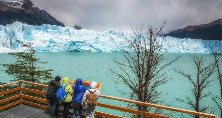 Des voyageurs en vestes colorées se tiennent sur une terrasse en bois surplombant le lac turquoise et l'imposant glacier Perito Moreno.