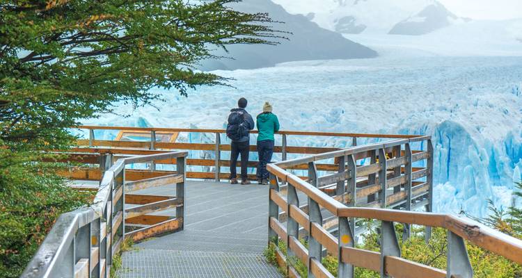 Un couple se tient sur une passerelle métallique entourée de feuillage, contemplant une vaste plaine glaciaire.
