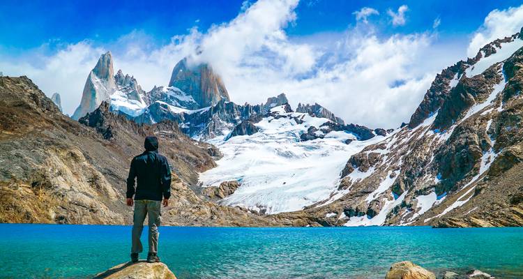 Un randonneur se tient à côté d'un lac glaciaire azur sous les flèches enneigées du Fitz Roy par ciel lumineux.
