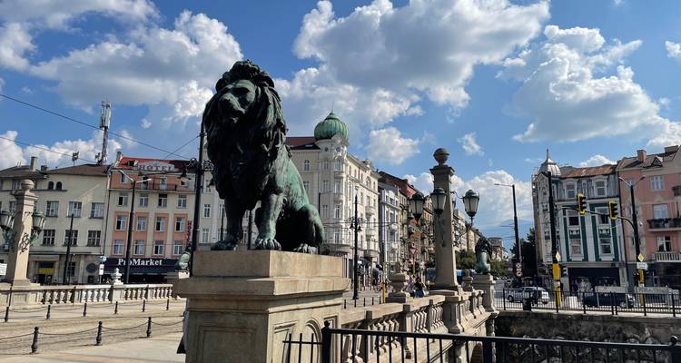 Bronze lion statue guards Sofia’s Lion Bridge with classic tram wires and bustling city buildings behind.