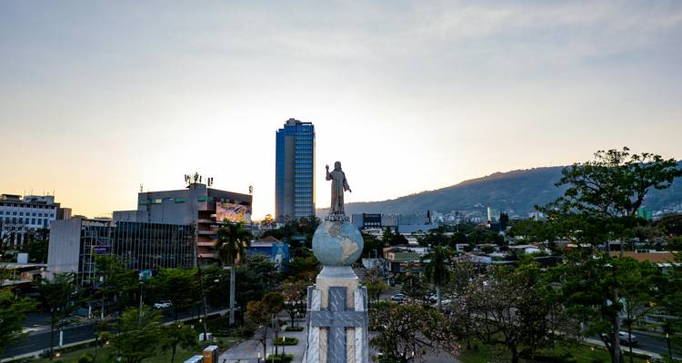 Estatua de Jesús de pie sobre un globo terráqueo con vista a un horizonte urbano moderno con montañas de fondo al atardecer.