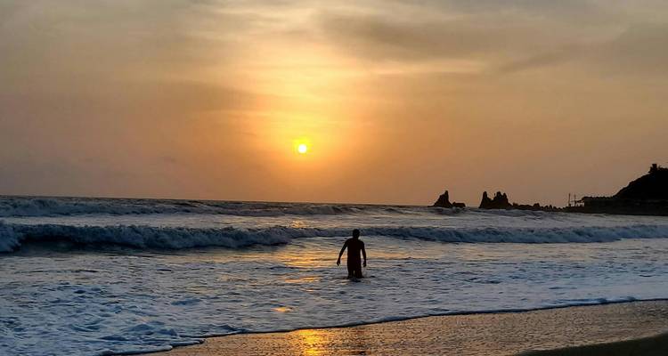 Silueta de un bañista solitario caminando en las olas mientras el sol se pone sobre una playa escarpada del Pacífico.