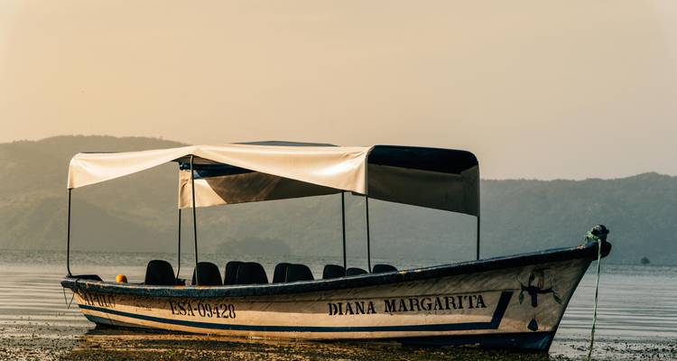 Un barco turístico cubierto flota en un lago tranquilo contra siluetas de montañas brumosas durante la hora dorada.