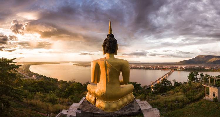 Goldene Buddha-Statue überblickt den Mekong-Fluss und die Stadt Pakse bei dramatischem Sonnenuntergang mit Sturmwolken.