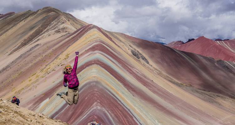 Excited hiker jumps for joy in front of Rainbow Mountain’s colourful layers under cloudy sky.