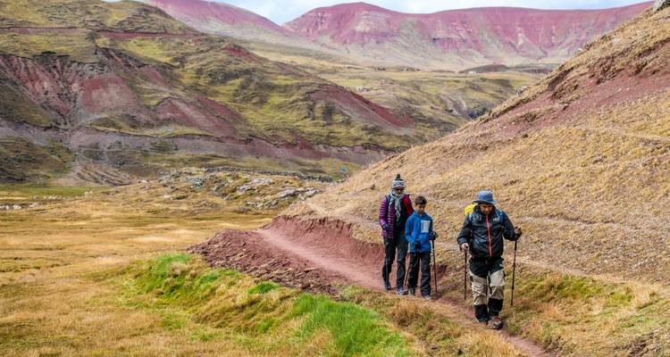 Small group of hikers follow dirt trail through colourful Andean valley flanked by rolling hills.