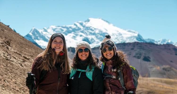 Three smiling trekkers pose with snow-covered Andean giants gleaming in bright sunshine behind.