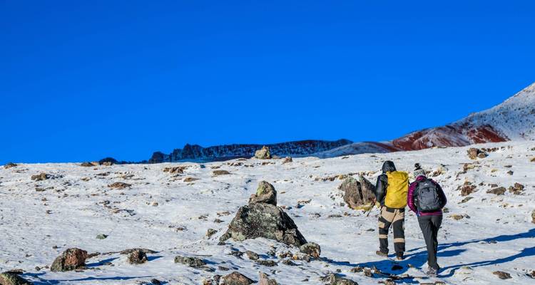 Two hikers trek across snow-dusted plateau beneath vivid blue sky towards distant red peaks.