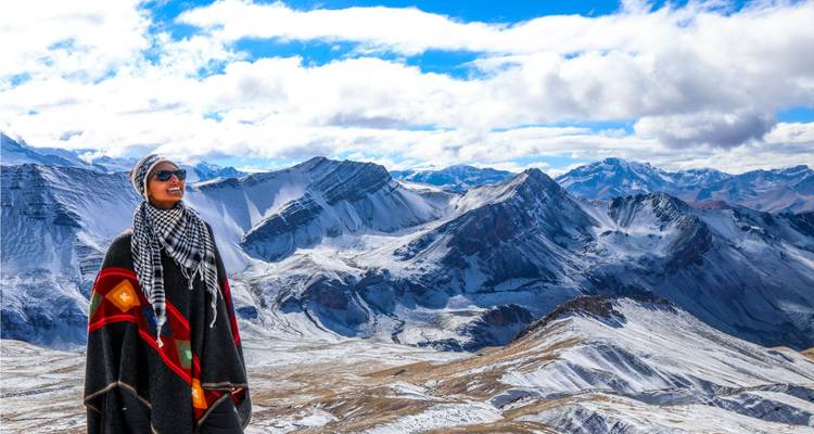 Traveller wrapped in Andean poncho smiles overlooking vast snowy mountain panorama under bright clouds.
