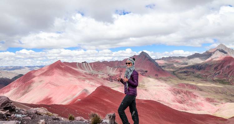 Traveller in cold-weather gear standing before vivid red valleys of Rainbow Mountain.