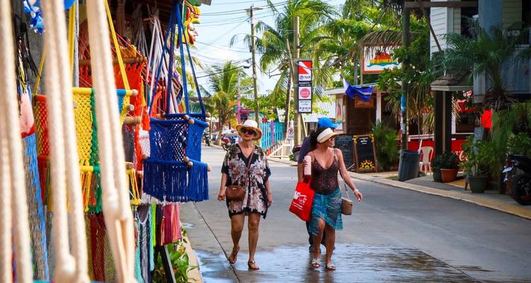 Two women carrying shopping bags stroll past colorful hammocks and signs on a lively tropical street.