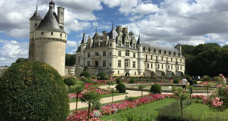 Elegant Château de Chenonceau with manicured flower gardens and moat under partly cloudy skies.
