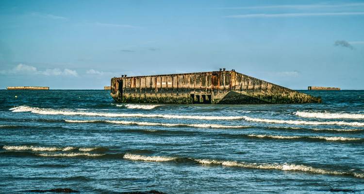 Overblijfselen van een Mulberry Harbour blok liggen in de golven voor een Normandische kust met kleine witte schuimkoppen onder een heldere hemel.