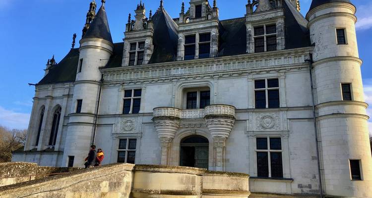 Groot Renaissance-stijl kasteel gevel met twin conische torens; een paar wandelt langs het stenen terras onder een heldere blauwe hemel.