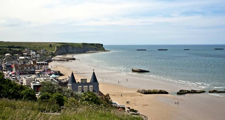 Verhoogd panorama van het dorp Arromanches-les-Bains, zandstrand met verspreide bezoekers, kliffen en overblijfselen van WOII-haven voor de kust.