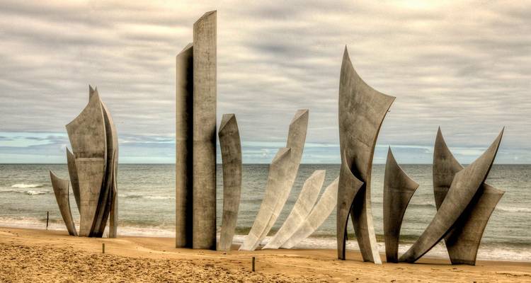 Opvallende roestvrijstalen sculptuur 'Les Braves' rijst op uit het zand op Omaha Beach tegen een gemoedvolle lucht en zachte branding.