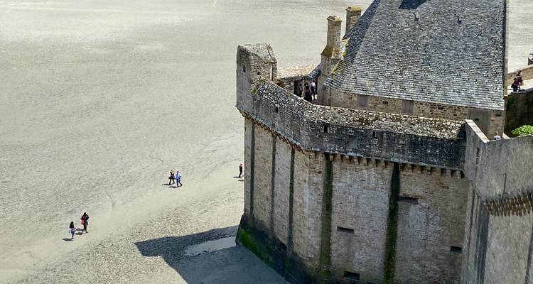 Stenen wallen van Mont Saint-Michel rijzen op boven uitgestrekte getijzanden waar kleine bezoekers ver beneden lopen.