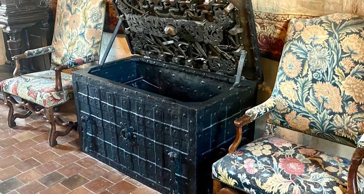 Antique iron strongbox flanked by richly upholstered Renaissance chairs on terracotta floor tiles inside a château room.