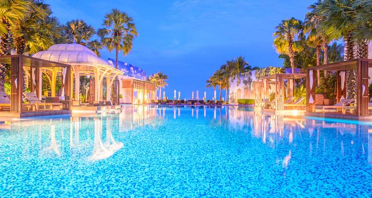 Piscine luxueuse de complexe hôtelier au crépuscule avec palmiers, cabanes et lumières brillantes reflétées dans l'eau.