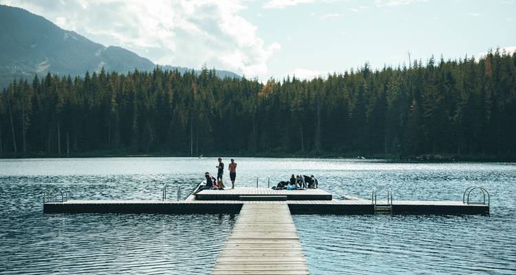 Wooden jetty extends into calm lake where people sit and relax, framed by dark forest