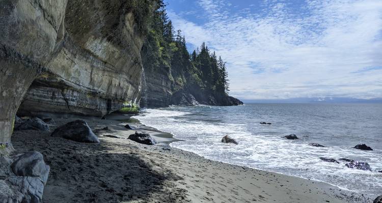 Rocky Pacific coastline with forested cliffs and gentle surf under a partly cloudy sky