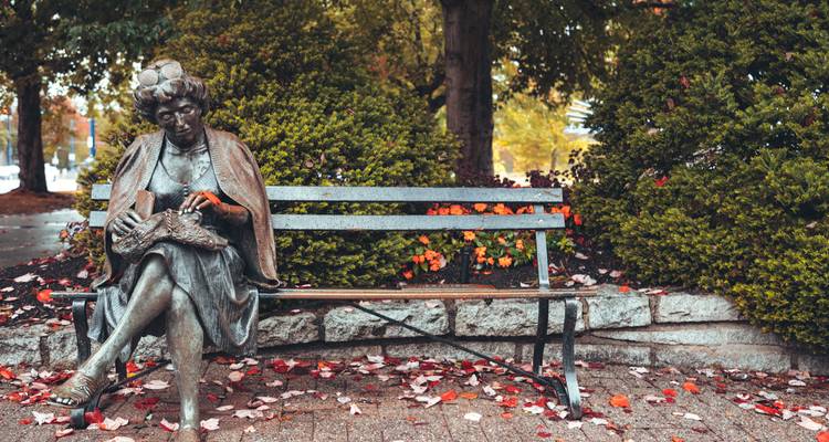 Bronze statue of a seated woman on a park bench surrounded by colourful autumn leaves and shrubs