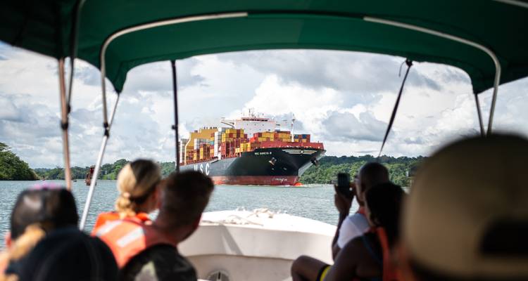 Small tour boat with passengers observing a large container ship transiting the Panama Canal amid lush green banks