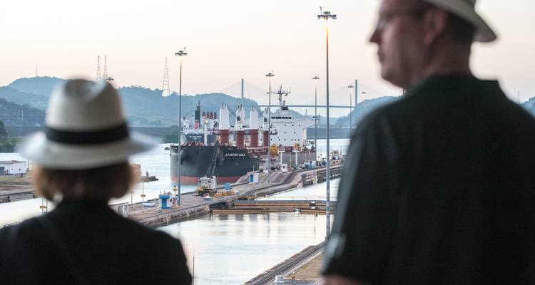 Two visitors in hats watch a cargo vessel pass through a lock of the Panama Canal at dusk