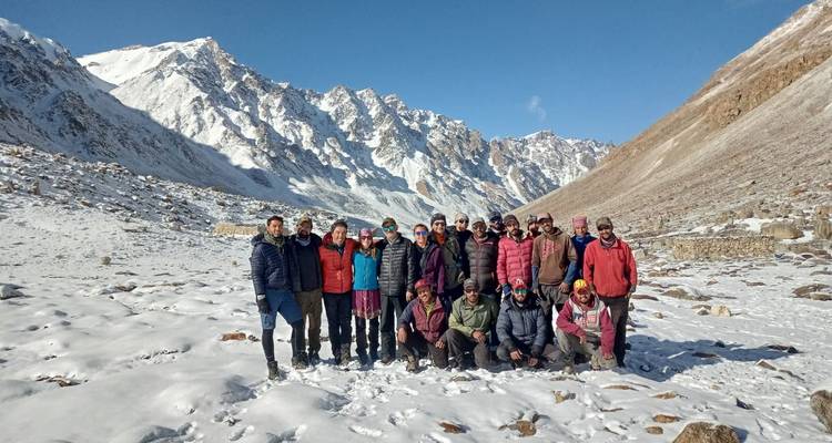 Gran grupo de senderismo posando en el suelo nevado del valle rodeado de picos blancos dentados del Himalaya bajo un cielo azul.