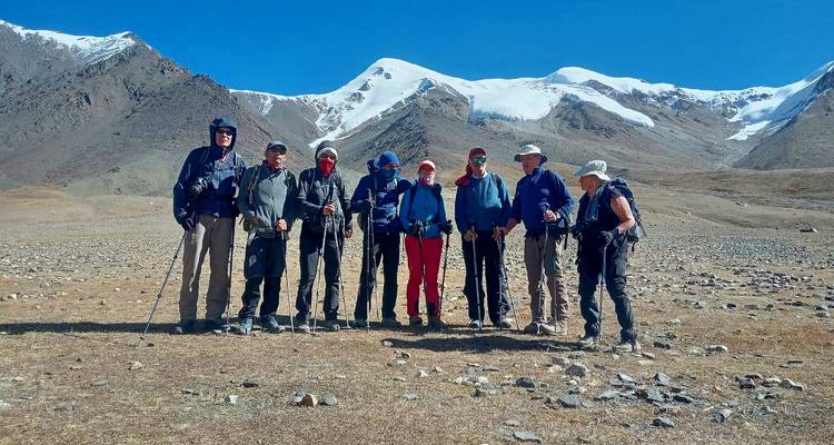 Pequeño grupo de excursionistas de pie en una meseta seca respaldada por crestas nevadas y cielo despejado.
