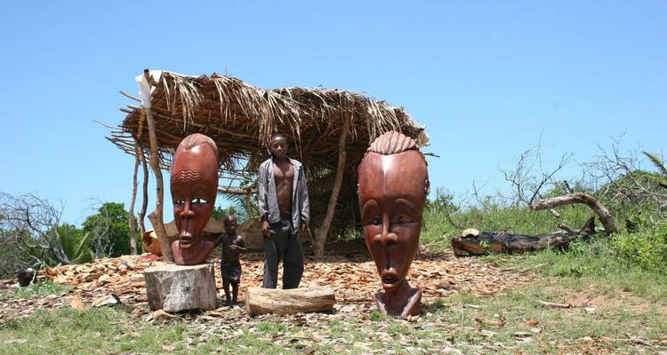 Local woodcarver and child stand beside two large carved wooden masks under a rustic palm-thatched shelter on grassy ground.