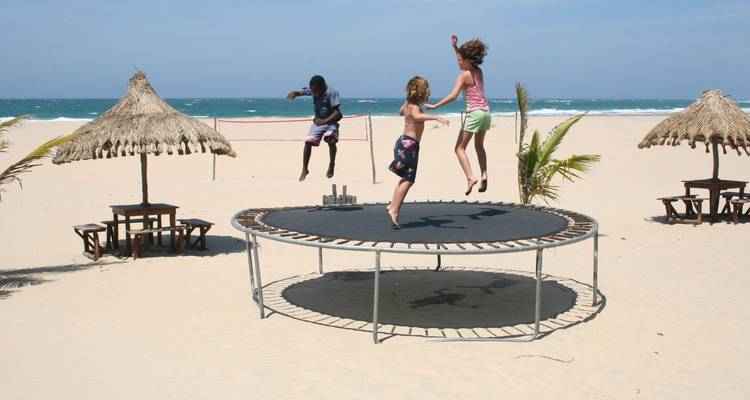 Drie kinderen springen op een trampoline die op een breed zandstrand staat met rieten parasols en blauwe oceaangolven op de achtergrond.