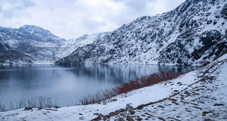 Icy alpine lake mirrors rugged snow-covered mountains and cloudy winter sky along a rocky trail.