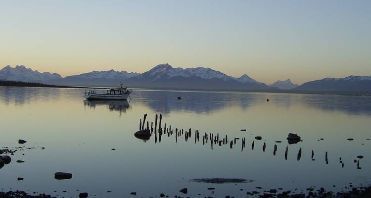 Kleine boot rust op spiegelkalme Patagonische fjord bij dageraad met gekartelde sneeuwtoppen in de verte.