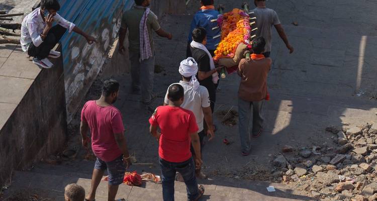 Des hommes portent un corps recouvert de fleurs le long de marches en pierre pendant une procession funéraire au bord de la rivière.