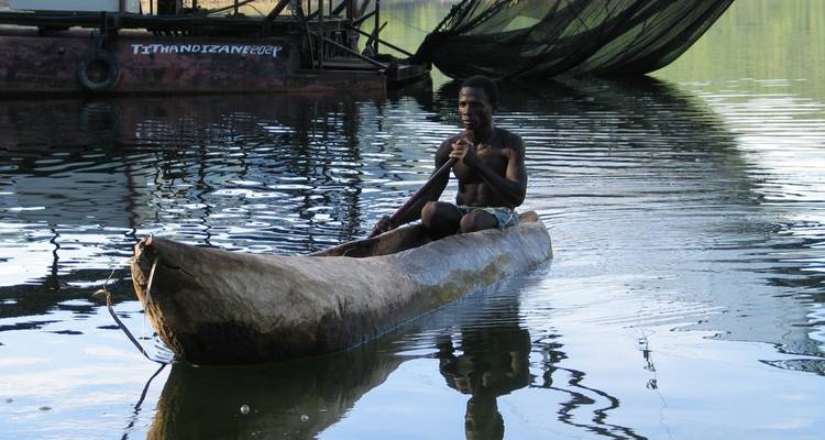 Man paddles a carved wooden canoe on calm reflective waters near nets and boats at sunset.