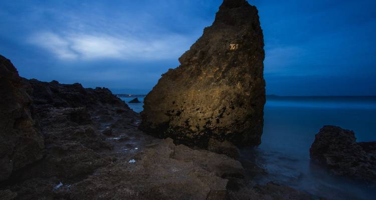 Tall jagged sea stack lit against a deep-blue twilight sky with smooth long-exposure ocean water around rocky shoreline.