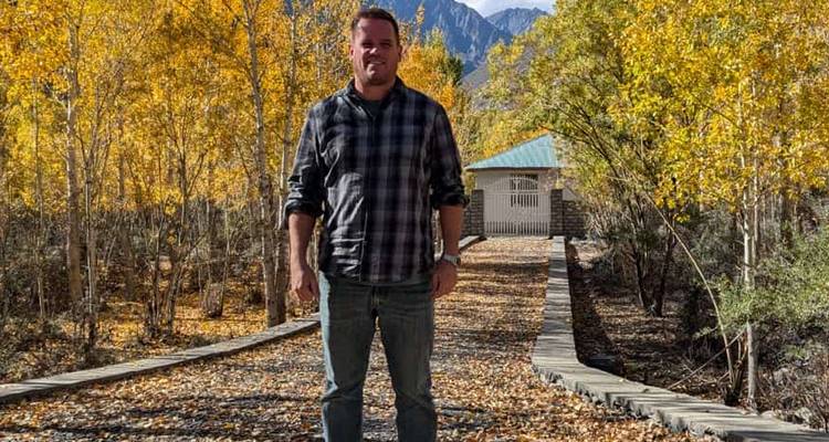 Un hombre está de pie en un sendero de madera cubierto de hojas entre álamos dorados de otoño con picos distantes.