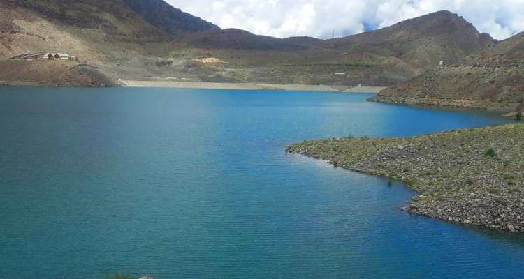 Un lago alpino turquesa se curva entre colinas rocosas áridas bajo un cielo nublado.