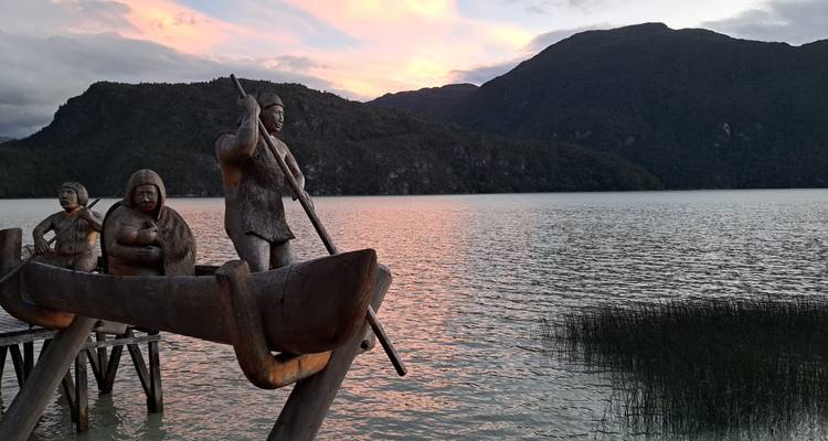 Escultura de madera de remeros indígenas en un muelle contempla un lago tranquilo al atardecer.