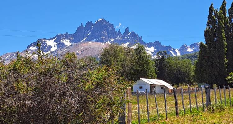 Pico de montaña escarpado cubierto de nieve que se alza detrás de una pequeña granja, cerca y prado en un día brillante y despejado.